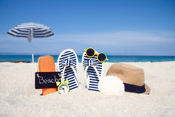 Obraz premium A Beach summer holiday banner background. Flip-flops and hat with a board and ball on the sand near the ocean. Summer accessories on the seashore. 