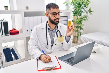 Young hispanic man doctor holding pills bottle writing on document at clinic