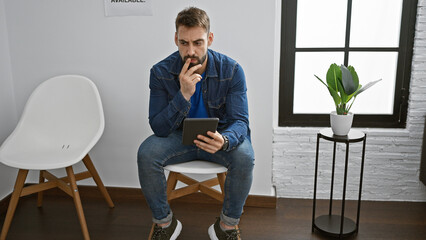 Young hispanic adult with a beard, deep in thought, sitting relaxed in the waiting room, using his...