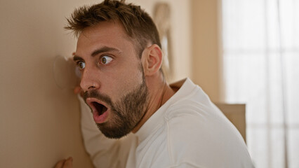 Curious young hispanic man surprised while eavesdropping through wall at home using a glass