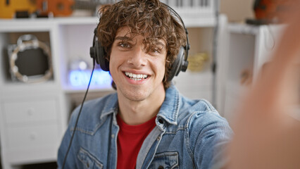 A smiling young man with curly hair wearing headphones and a denim jacket in a modern music studio.