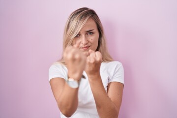 Young blonde woman standing over pink background ready to fight with fist defense gesture, angry...