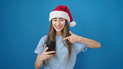 Young beautiful hispanic woman wearing christmas hat pointing to smartphone over isolated blue background