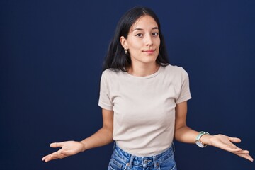 Young hispanic woman standing over blue background clueless and confused with open arms, no idea...
