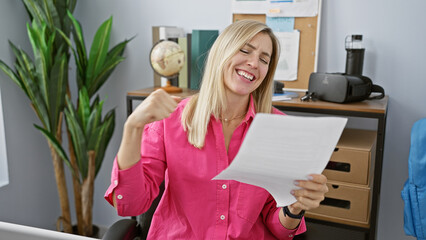 A cheerful blonde woman wearing a pink shirt exults while reading a paper in a modern office setting.