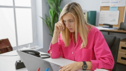 A focused young blonde woman works thoughtfully at her desk in a modern office setting.