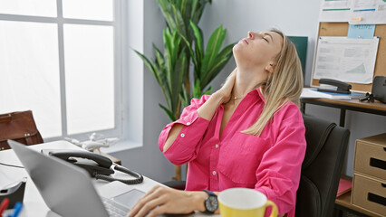 A young, blonde woman in a pink shirt feeling neck pain at her office desk, exuding a mix of professionalism and discomfort.