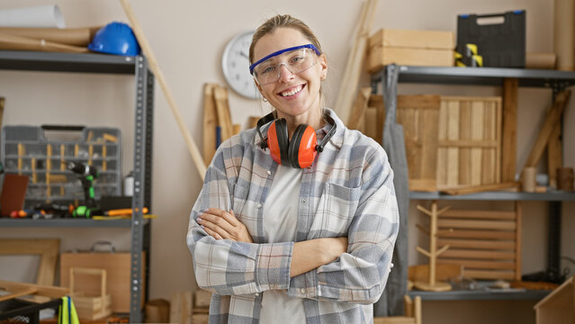 Confident young woman with arms crossed wearing safety goggles and ear protection in a well-organized carpentry workshop