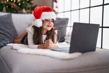 Adorable hispanic girl having video call lying on sofa by christmas tree at home