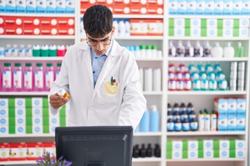 Young hispanic man pharmacist using computer holding pills bottle at pharmacy