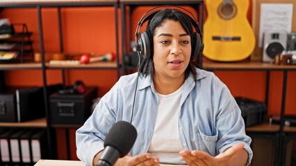 Hispanic woman podcasting in a music studio with headphones, microphone, and guitar background.