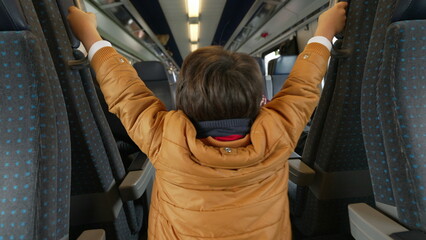 Back of Child playing inside train holding into passenger seats standing in corridor at high-speed...