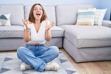 Young caucasian woman sitting on the floor at the living room celebrating crazy and amazed for...