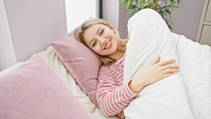 A smiling young woman lies comfortably in bed with pink pillows, offering a warm, welcoming home feel.