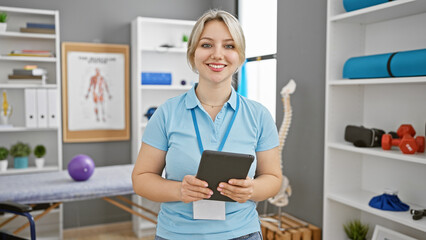 Confident young woman clinician with blonde hair holding a tablet in a well-equipped rehab clinic interior.