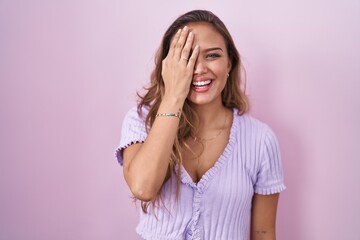 Young hispanic woman standing over pink background covering one eye with hand, confident smile on face and surprise emotion. - obrazy, fototapety, plakaty