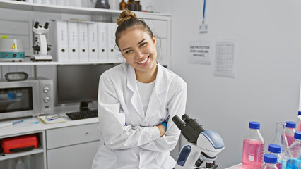 Smiling young hispanic scientist woman, beautiful and professional, sitting in the lab with arms crossed, working confidently with a microscope to discover medical breakthroughs. - obrazy, fototapety, plakaty
