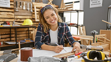 Young, beautiful hispanic woman - a passionate carpenter, sitting at her carpentry workshop, smiling behind safety glasses, happily taking construction notes for her bespoke furniture design project. - obrazy, fototapety, plakaty