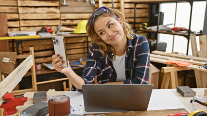 Young, beautiful hispanic woman carpenter captures her online woodworking business on laptop while taking a smiling selfie at carpentry workshop