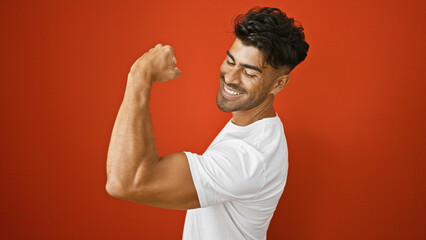 Handsome young hispanic man flexing muscles with a smile against a red wall background - obrazy, fototapety, plakaty