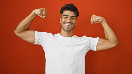 A cheerful young hispanic man flexes his muscles confidently against an isolated red background,...