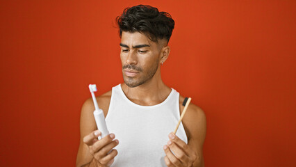 Handsome hispanic man confused between manual and electric toothbrushes against a red wall