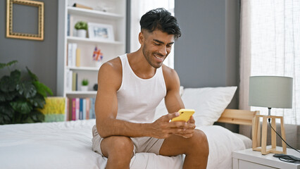 Obraz na płótnie Canvas Smiling hispanic man using smartphone in modern bedroom setting.