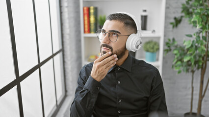 Pensive young man with beard and glasses wearing headphones in modern indoor office setting.