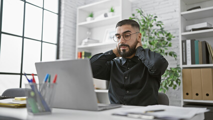 A young bearded man wearing glasses feels stressed while working on a laptop in a modern office.