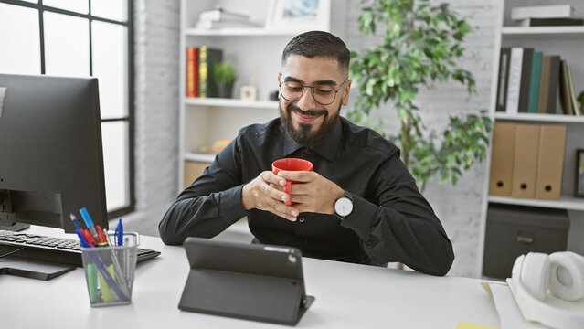 Smiling bearded man with mug at modern office desk using tablet