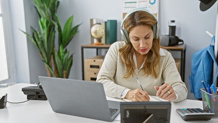 A focused hispanic woman wearing headphones takes notes while working with a laptop in a modern...