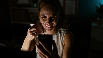 Smiling caucasian woman holding coffee mug and smartphone in a dimly lit room