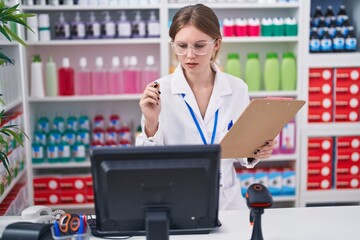 Young blonde woman pharmacist using computer reading document at pharmacy