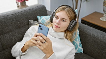 Blonde woman with headphones uses smartphone on couch, portraying a casual indoor setting, creating a relatable leisure scene.