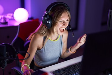 Young blonde woman streamer playing video game with winner expression at gaming room