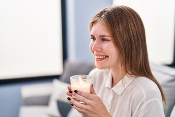 Young blonde woman sitting on sofa smelling aromatic candle at home