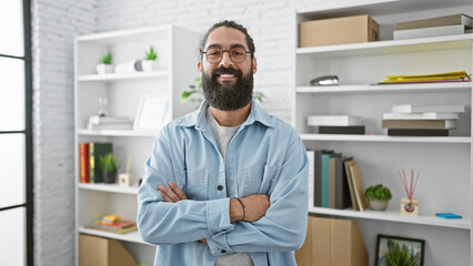 A confident hispanic man with a beard standing in a modern office setting, arms crossed and smiling.