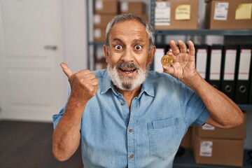 Cheerful senior man showing thumbs up, holding a bitcoin coin in office, smiling wide with open...