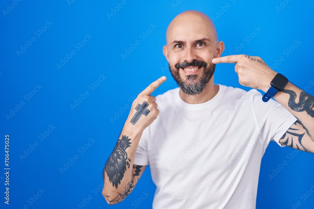 Canvas Prints Hispanic man with tattoos standing over blue background smiling cheerful showing and pointing with fingers teeth and mouth. dental health concept.