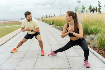 Man and woman doing stretching exercises outdoors, in sync and smiling, with a city skyline in the background.