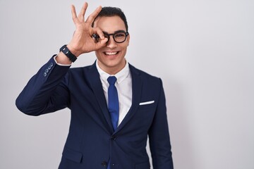 Young hispanic man wearing suit and tie doing ok gesture with hand smiling, eye looking through fingers with happy face.
