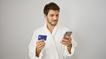 Handsome hispanic man in white bathrobe holding credit card and smartphone against isolated background.