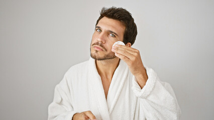 Handsome young man in white bathrobe applying facial skincare on isolated background