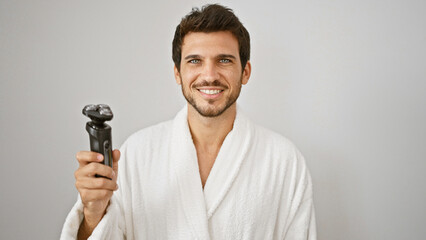 Handsome hispanic man with beard smiling in white robe holding electric shaver against isolated...