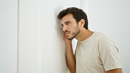 Hispanic young man with a beard casually leaning on a white interior door at home, looking pensive.