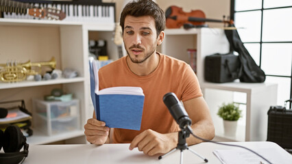 Handsome hispanic man reading book indoors at a podcasting studio microphone