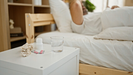 Sick person resting in bed with medicine, water, and tissues on nightstand in a cozy bedroom.