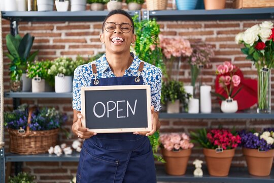 African american woman working at florist holding open sign sticking tongue out happy with funny expression. - Powered by Adobe