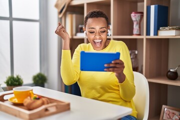 African american woman using touchpad sitting on the table at breakfast screaming proud, celebrating victory and success very excited with raised arm