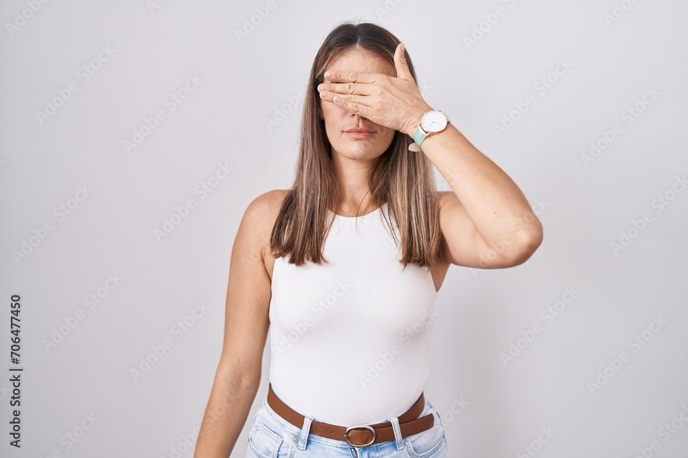 Sticker hispanic young woman standing over white background covering eyes with hand, looking serious and sad